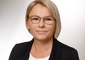 Porträtfoto einer erwachsenen Person mit kurzen blonden Haaren und runder Brille. Die Person trägt schwarzen Blazer über weißer Bluse, blickt direkt in die Kamera; neutraler Hintergrund, ruhiger, professioneller Ausdruck.