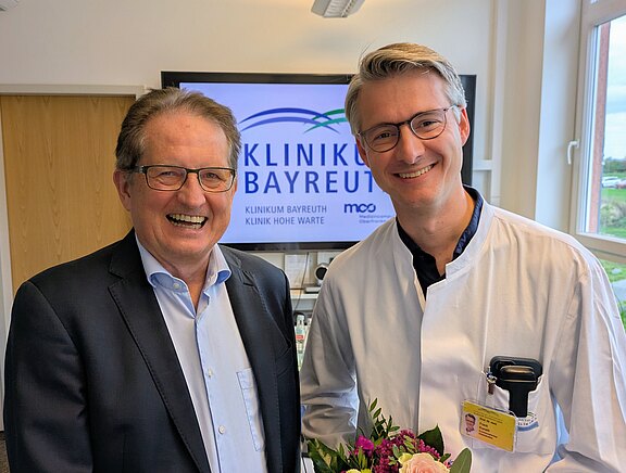 Zwei Männer stehen lächelnd in einem Raum, der mit einem Bildschirm im Hintergrund ausgestattet ist, auf dem das Logo der Klinik Bayreuth zu sehen ist. Der Mann links trägt eine Brille und einen Anzug, der Mann rechts eine Arztkittel und hält einen Blumenstrauß.