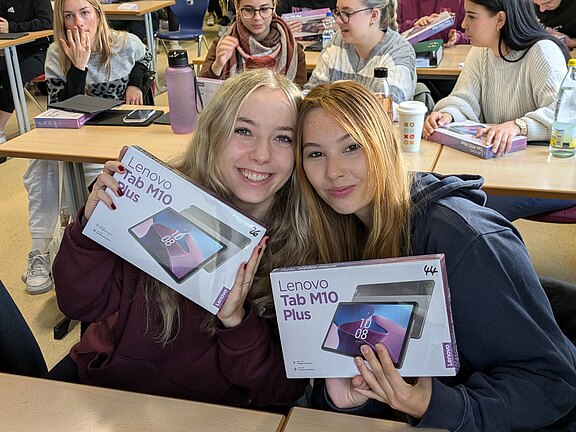 Zwei Schülerinnen sitzen im Klassenraum und halten lächelnd je eine Schachtel mit „Lenovo Tab M10 Plus“ in die Kamera. Im Hintergrund weitere Lernende. Botschaft: Freude über neue Tablets für den Unterricht.
