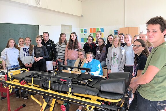 Schulklasse versammelt in einem Klassenraum um eine gelbe Rettungstrage; sie schauen zur Kamera und wirken bei einer Erste-Hilfe- oder Sanitätsvorführung.