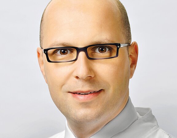 Porträt eines lächelnden Mannes mit Glatze und dunkler Brille vor neutralem Hintergrund. Nahaufnahme des Gesichts, direkter Blick in die Kamera, seriöser Ausdruck, Business-Hemd.