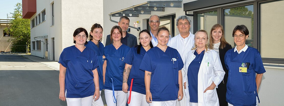Gruppe von Pflegekräften und Ärztinnen/Ärzten in blauen Kitteln und weißen Mänteln steht lächelnd vor einem modernen Klinikgebäude und blickt in die Kamera.