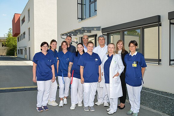 Gruppe von Pflegekräften und Ärztinnen/Ärzten in blauen Kitteln und weißen Mänteln steht lächelnd vor einem modernen Klinikgebäude und blickt in die Kamera.