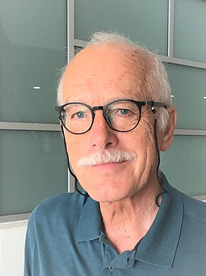 Porträt eines älteren Mannes mit weißem Schnurrbart und runder Brille mit Band. Er trägt ein blaugrünes Polohemd, blickt zur Seite. Neutraler Innenhintergrund.