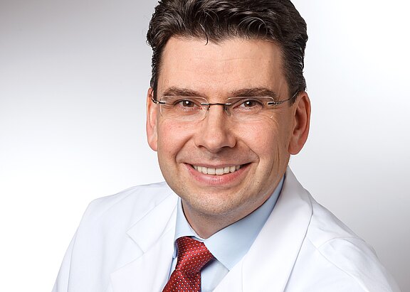 Porträtfoto eines lächelnden Mannes mit Brille im weißen Kittel und roter Krawatte vor neutralem Hintergrund.