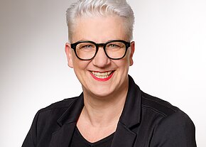 Porträtfoto einer lächelnden Person mit kurzem weißem Haar, schwarzer Brille und rotem Lippenstift, in schwarzer Jacke vor hellem, neutralem Hintergrund; Blick frontal in die Kamera.