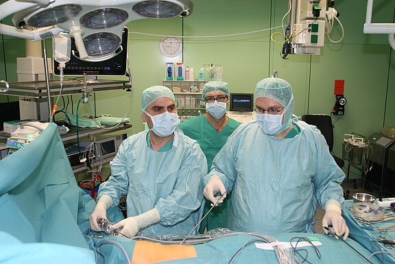 Mehrere Chirurgen in OP-Kleidung führen eine minimalinvasive Operation durch. Sie arbeiten konzentriert an einem Patienten, der unter sterilen Tüchern liegt, nutzen laparoskopische Instrumente und schauen auf Monitore. Helles OP-Licht und medizinische Geräte umgeben den Tisch.