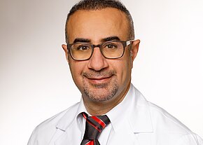 Ein freundlicher männlicher Arzt mit Brille und kurzem, grauem Bart lächelt in die Kamera. Er trägt einen weißen Laborkittel und ein gestreiftes, schwarzes und rotes Hemd mit Krawatte. Der Hintergrund ist hell und neutral.