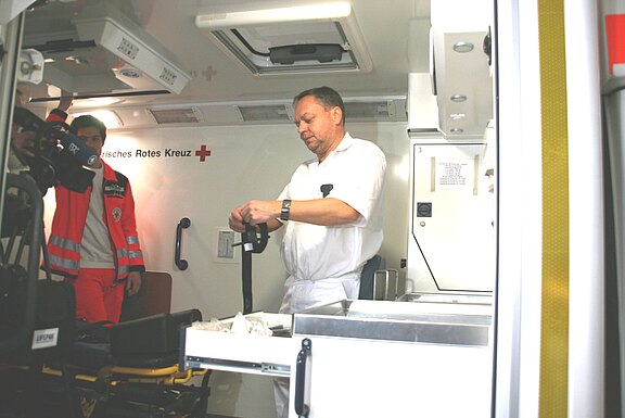 Innenraum eines Rettungswagens: Ein medizinischer Mitarbeiter in Weiß bereitet ein Gerät an der Arbeitsfläche vor. Daneben steht eine Person im roten Einsatzanzug des Deutschen Roten Kreuzes. Behandlungsbereich mit Trage und Schubladen sichtbar.