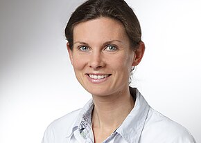 Dr. med. Katharina Link Porträtfoto einer lächelnden Person mit hellen Augen und zusammengebundenen Haaren, in weißem Kittel, vor neutralem, hellem Hintergrund. Fokus auf freundlichem Gesichtsausdruck.