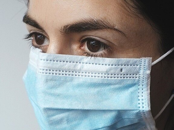Nahaufnahme einer Person mit blauem medizinischen Mund-Nasen-Schutz; der Blick geht zur Seite, die Augen sind deutlich sichtbar und betonen das Thema Gesundheitsschutz.