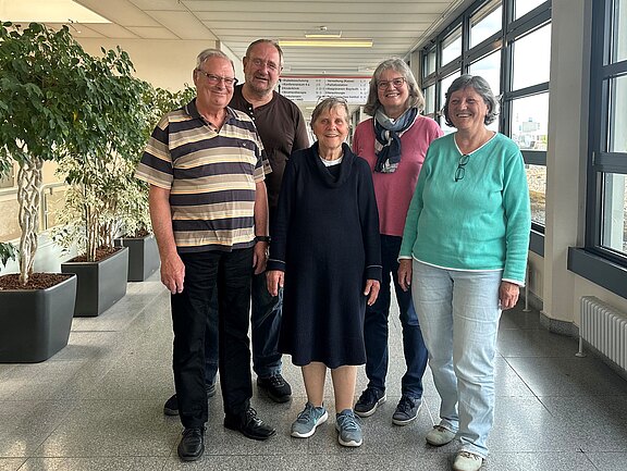 Fünf Personen stehen in einem hellen Raum mit großen Fenstern. Sie lächeln und tragen legere Kleidung. Im Hintergrund sind Pflanzen in Töpfen sichtbar. Die Personen scheinen sich wohlzufühlen und sind freundlich zueinander.