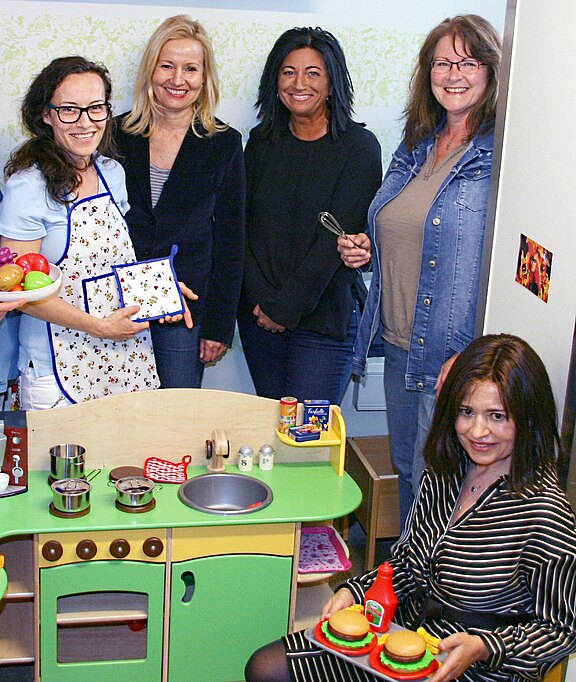 Fünf Frauen stehen und hocken um eine bunte Kinderküche, lächeln in die Kamera. Eine trägt eine Schürze; eine andere hält eine Zange; vorne präsentiert jemand zwei Spiel-Burger mit Ketchup. Auf der Arbeitsfläche liegen Töpfe, Pfannen und Spiellebensmittel.