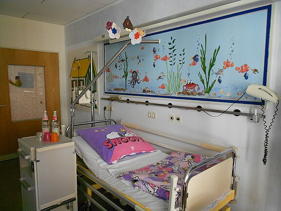 Kinderkrankenhauszimmer mit Einzelbett und bunter Bettwäsche, daneben Nachttisch mit Getränken. Über dem Bett hängt ein Trapezbügel. Die Wand zeigt ein großes Unterwasserbild mit Fischen und Pflanzen, das den Raum freundlich wirken lässt.