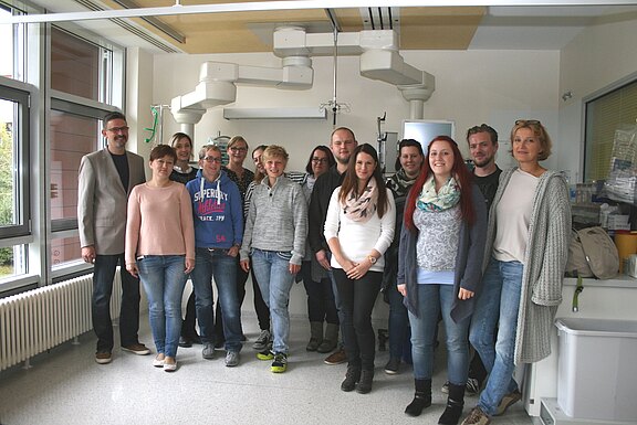 Eine Gruppe von Erwachsenen posiert für ein Gruppenfoto in einem hellen, klinisch wirkenden Raum mit medizinischer Ausstattung. Alle schauen zur Kamera; die Stimmung ist freundlich.