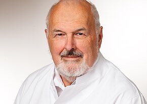 Porträt eines älteren Mannes mit weißem Haar und Bart, in weißem Kittel, freundlich in die Kamera blickend vor neutralem Hintergrund.