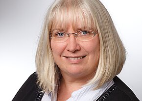 Porträtfoto: hellblonde Frau mit schulterlangem Haar und randloser Brille, lächelt freundlich. Sie trägt weiße Bluse unter dunkler Jacke. Neutraler, heller Hintergrund.