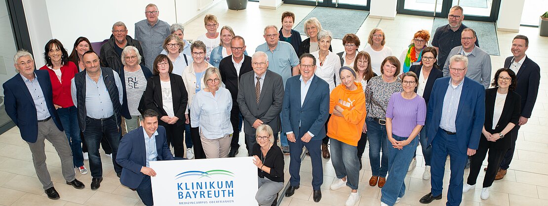 Gruppenfoto von Mitarbeitenden in einer hellen Eingangshalle: Rund 30 Erwachsene stehen lächelnd zusammen; vorn halten zwei ein Schild mit der Aufschrift „Klinikum Bayreuth“. Die Szene vermittelt Teamzusammenhalt bzw. ein gemeinsames Treffen.