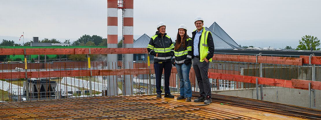 Drei Personen mit Schutzhelmen und Warnwesten stehen auf einer Baustelle auf einer bewehrten Betonplatte und schauen in die Kamera.