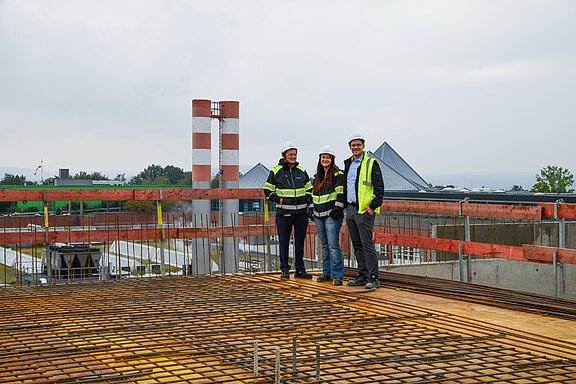 Drei Personen mit Schutzhelmen und Warnwesten stehen auf einer Baustelle auf einer bewehrten Betonplatte und schauen in die Kamera.