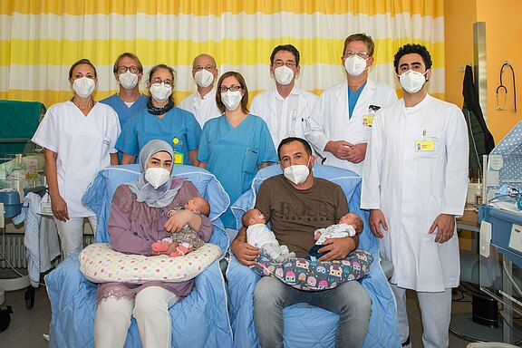 Im Krankenhaus sitzen zwei Eltern auf Sesseln und halten ihre neugeborenen Zwillinge. Hinter ihnen steht ein Team aus Ärztinnen, Ärzten und Pflegekräften in Kitteln. Alle tragen Schutzmasken. Die Szene wirkt wie ein offizielles Gruppenfoto nach einer Geburt.