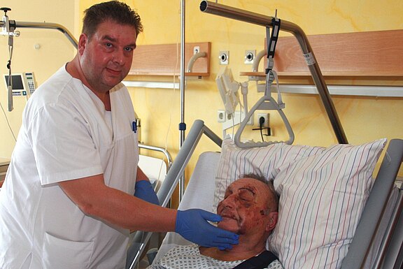 Pflegekraft in weißer Kleidung und blauen Handschuhen betreut einen älteren Mann mit Gesichtsverletzungen im Krankenhausbett. Sie hält sanft sein Kinn; der Patient liegt halb aufgerichtet und wirkt erschöpft, aber ruhig.