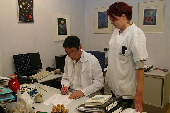 Ein Arzt in weißem Kittel sitzt am Schreibtisch und unterschreibt Unterlagen. Neben ihm steht eine Pflegekraft, die auf die Papiere schaut. Beide wirken konzentriert, als würden sie Patientendaten oder einen Bericht durchgehen.