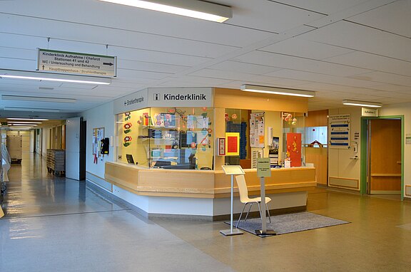 Heller Krankenhausflur mit runder Empfangstheke der Kinderklinik. Der Schalter hat Glasfenster, dahinter ein Arbeitsbereich, dekoriert mit bunten Kinderzeichnungen. Ein Wegweiser hängt an der Decke, Stühle stehen vor dem Tresen, Türen führen zu Behandlungsräumen.