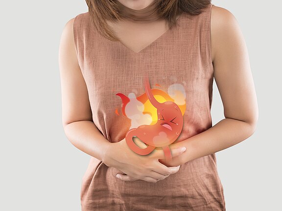 Person hält sich den Bauch; über dem Magen ist eine brennende, schwitzende Cartoon-Darstellung eingeblendet. Bild symbolisiert Sodbrennen, Magenschmerzen oder Verdauungsbeschwerden.
