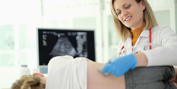 Ärztin führt bei einer liegenden Person einen Bauch-Ultraschall durch. Sie setzt den Schallkopf am Unterbauch an; auf dem Monitor ist das resultierende Ultraschallbild zu sehen.