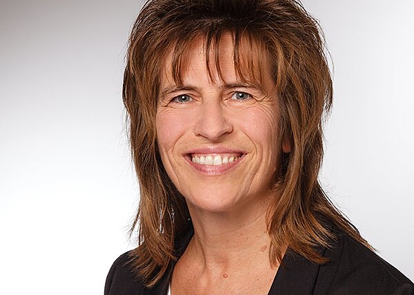 Porträtfoto einer lächelnden Frau mittleren Alters mit braunen, schulterlangen Haaren; sie trägt ein dunkles Jackett über einem weißen Oberteil. Neutraler, hellgrauer Hintergrund.