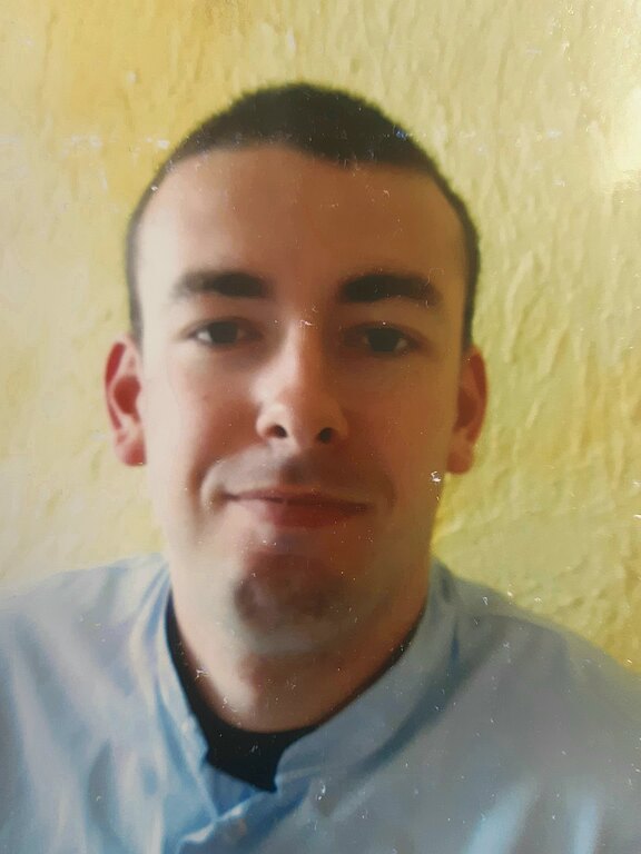 Nahaufnahme einer Person mit kurzen dunklen Haaren vor gelblicher Wand. Die Person blickt direkt in die Kamera und lächelt leicht. Ein heller Kragen ist sichtbar.