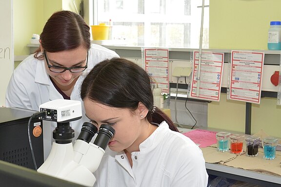 Zwei Frauen in Laborkitteln arbeiten in einem Labor. Eine Frau schaut durch ein Mikroskop, während die andere sie beobachtet und unterstützt. Im Hintergrund sind verschiedene Glasbehälter mit farbigen Flüssigkeiten und Informationsblätter zu sehen.