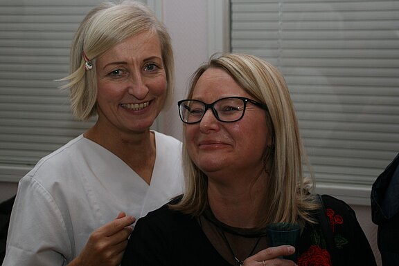 Zwei erwachsene Frauen stehen eng beieinander und lächeln in die Kamera. Die linke trägt weiße Kleidung, die rechte eine Brille und hält einen Becher. Die Szene wirkt freundlich und ungezwungen in einem Innenraum.