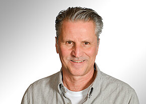 Porträt eines lächelnden Mannes mittleren Alters mit grauen, zurückgekämmten Haaren. Er trägt ein helles Hemd über einem weißen T‑Shirt. Neutraler Hintergrund. Freundlicher Gesichtsausdruck, Blick in die Kamera.