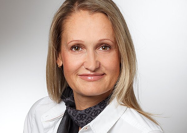 Porträt einer lächelnden Frau mit langen blonden Haaren. Sie trägt eine weiße Bluse und ein dunkles Halstuch. Neutrale Studioaufnahme vor hellem Hintergrund.