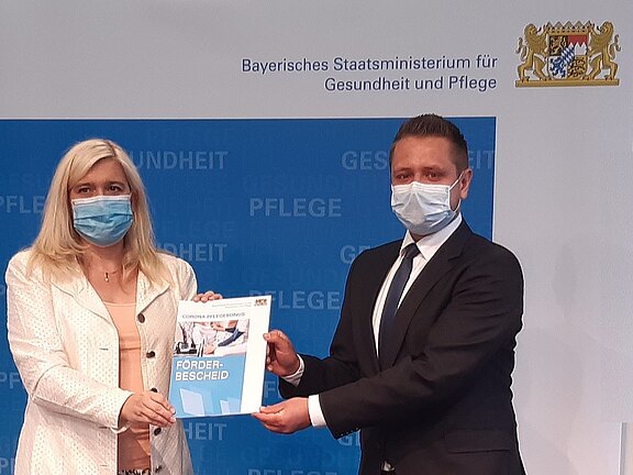 Zwei Personen mit Mund-Nasen-Schutz in formeller Kleidung präsentieren gemeinsam einen Förderbescheid. Sie stehen vor einer Wand mit dem Logo und Schriftzug des Bayerischen Staatsministeriums für Gesundheit und Pflege.