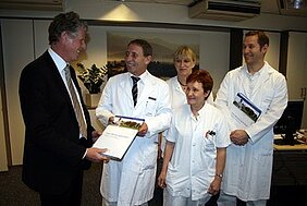 Eine Gruppe von vier Personen in weißen Arztkitteln steht in einem Büro. Eine Person überreicht einem anderen eine Urkunde oder ein Zertifikat. Alle wirken erfreut und diskutieren miteinander. Das Bild vermittelt eine Atmosphäre der Anerkennung und Zusammenarbeit im medizinischen Bereich.