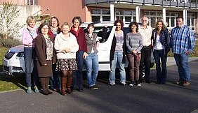 Eine Gruppe von zehn Personen steht lächelnd vor einem weißen Auto, vor einem Gebäude. Sie sind unterschiedlich gekleidet und scheinen sich gut zu verstehen. Die Szene vermittelt ein Gefühl von Gemeinschaft und Freude.