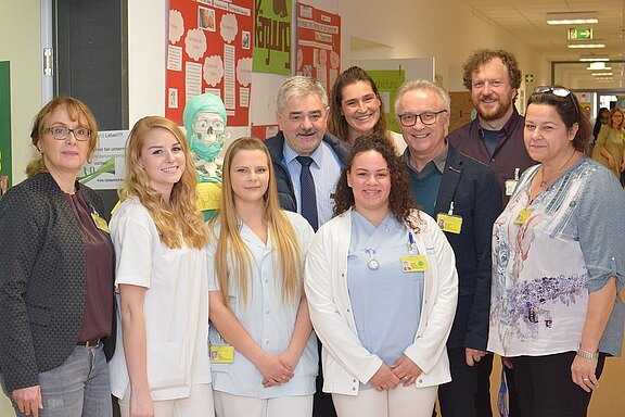 Eine Gruppe von Fachleuten steht im Flur eines Gesundheitszentrums. Sie tragen Arbeitskleidung und lächeln freundlich in die Kamera. Im Hintergrund sind Lehrmaterialien und ein Modell eines Skeletts sichtbar, das in medizinischen Kontext stellt.