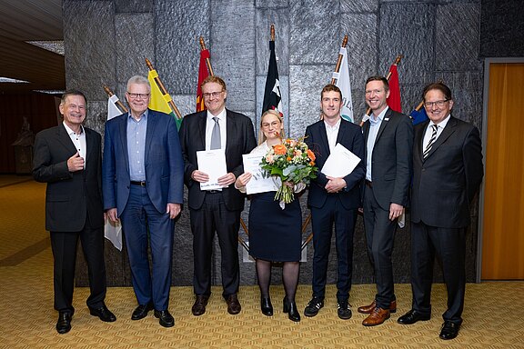 Eine Gruppe von sieben Personen steht in einem Raum, von denen einige Urkunden und eine Blumenstrauß in der Hand halten. Im Hintergrund sind Flaggen zu sehen, was auf eine offizielle Veranstaltung hinweist. Alle wirken erfreut und posieren für ein Foto.