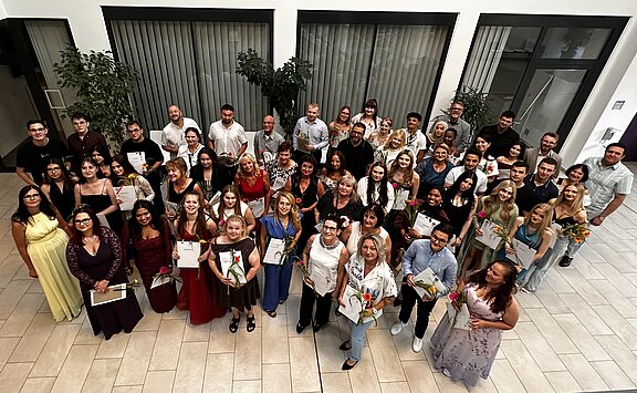 Großes Gruppenfoto in einem hellen Innenraum: zahlreiche Menschen in festlicher Kleidung stehen eng zusammen, viele halten Urkunden und Blumen. Alle blicken lächelnd nach oben in die Kamera. Der Moment wirkt wie eine Abschlussfeier oder Preisverleihung.