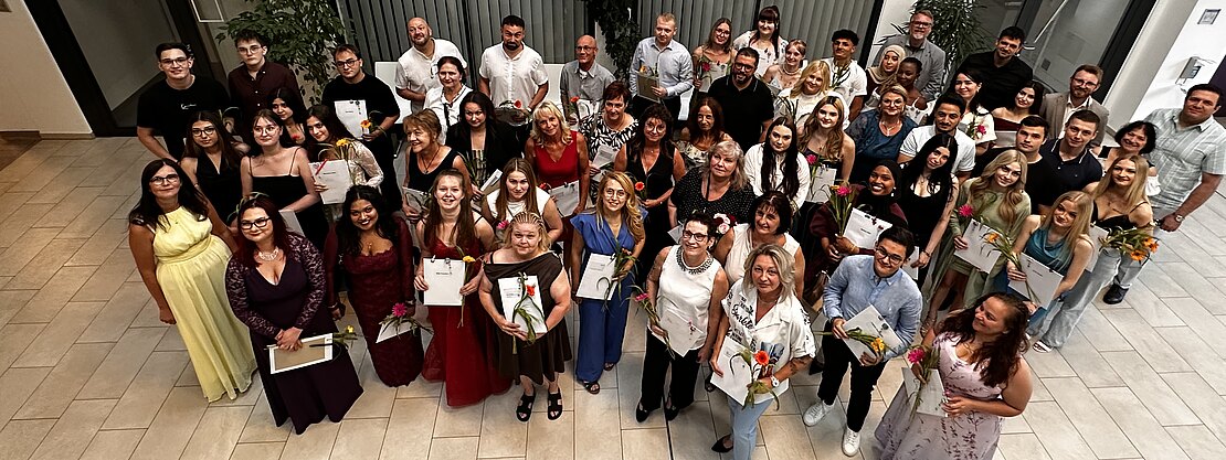 Großes Gruppenfoto in einem hellen Innenraum: zahlreiche Menschen in festlicher Kleidung stehen eng zusammen, viele halten Urkunden und Blumen. Alle blicken lächelnd nach oben in die Kamera. Der Moment wirkt wie eine Abschlussfeier oder Preisverleihung.