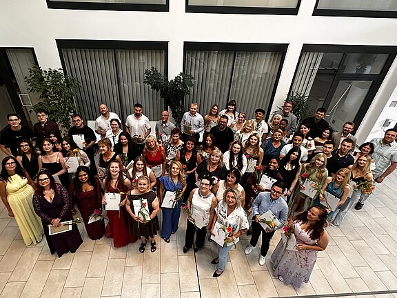 Großes Gruppenfoto in einem hellen Innenraum: zahlreiche Menschen in festlicher Kleidung stehen eng zusammen, viele halten Urkunden und Blumen. Alle blicken lächelnd nach oben in die Kamera. Der Moment wirkt wie eine Abschlussfeier oder Preisverleihung.