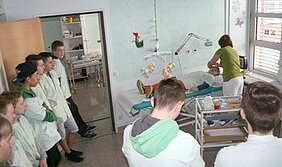 Eine Gruppe von Jugendlichen beobachtet einen medizinischen Eingriff oder eine Behandlung in einem Krankenhauszimmer. Eine Fachkraft kümmert sich um einen Patienten, der auf einem Krankenbett liegt. Die Umgebung ist sauber und praktisch gestaltet.