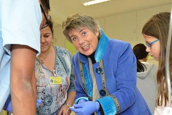 Eine lächelnde Frau in einem blauen Mantel interagiert mit mehreren Personen, während sie eine medizinische oder wissenschaftliche Aktivität durchführt. Sie trägt Einmalhandschuhe und scheint etwas zu erklären oder zu demonstrieren. Die Umgebung wirkt freundlich und informell.