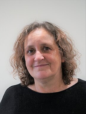 Susanne Dietze Nahaufnahme eines Kopf-Schulter-Porträts einer mittelalten Frau mit lockigem, schulterlangem braunem Haar. Sie trägt einen schwarzen Pullover, lächelt leicht und blickt direkt in die Kamera vor neutralem Hintergrund.
