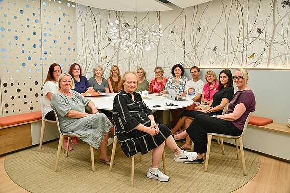 Vierzehn Erwachsene sitzen um einen runden Konferenztisch in einem hellen, modernen Raum und blicken zur Kamera. Sie wirken als Team in einer Besprechung oder Gruppenfoto-Situation.