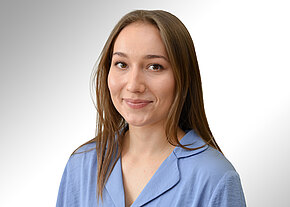 Porträtfoto: Eine junge Frau mit langem braunem Haar lächelt freundlich. Sie trägt ein hellblaues Oberteil mit Kragen. Neutraler, heller Hintergrund; Fokus auf freundlichem Ausdruck und professionellem Erscheinungsbild.