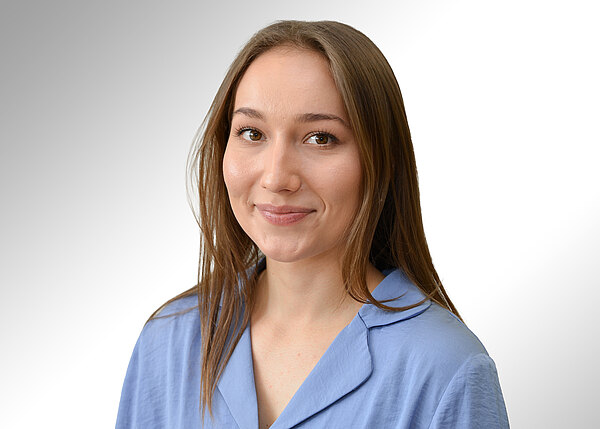 Porträtfoto: Eine junge Frau mit langem braunem Haar lächelt freundlich. Sie trägt ein hellblaues Oberteil mit Kragen. Neutraler, heller Hintergrund; Fokus auf freundlichem Ausdruck und professionellem Erscheinungsbild.